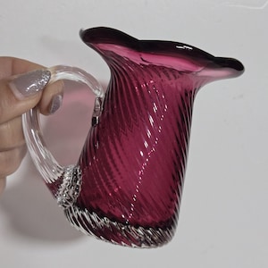 May include: A small, hand-blown glass pitcher in a deep magenta hue. The pitcher features a clear, curved handle and a ruffled rim. The body of the pitcher has a textured, swirled design. The base of the pitcher has a textured, swirled design.
