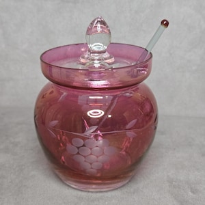 Könnte beinhalten: Ein vintage, rubinfarbenes Glasgefäß mit Deckel und einem kleinen Glaslöffel. Das Gefäß hat einen runden Körper mit geätzten Trauben- und Blattmotiven. Der Deckel hat einen Knauf aus klarem Glas. Der Löffel hat eine rote Spitze.