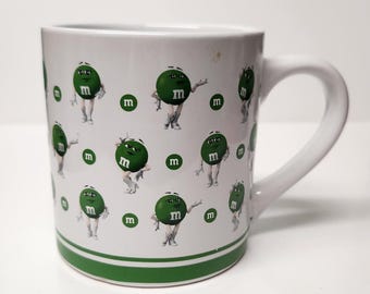 May include: White ceramic mug with a green rim and a pattern of green M&M's with faces on a white background.
