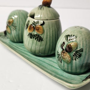 Puede incluir: Un juego de tres saleros y pimenteros de cerámica verde con diseños de piñas y una bandeja a juego. Los saleros tienen un acabado esmaltado y están decorados con piñas marrones y amarillas.