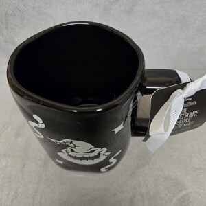 May include: A black ceramic mug featuring a white graphic of the character Oogie Boogie from The Nightmare Before Christmas. The mug has a glossy finish, a wide handle, and a white ribbon attached to a tag that reads Disney Tim Burton's The Nightmare Before Christmas.