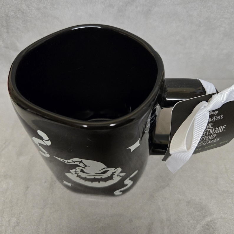 May include: A black ceramic mug featuring a white graphic of the character Oogie Boogie from The Nightmare Before Christmas. The mug has a glossy finish, a wide handle, and a white ribbon attached to a tag that reads Disney Tim Burton's The Nightmare Before Christmas.