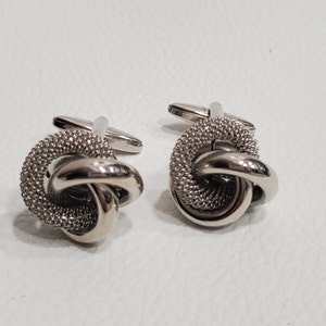 Könnte beinhalten: Ein Paar silberne Manschettenknöpfe mit einem Knotendesign. Die Knoten sind strukturiert und haben eine glänzende Oberfläche.