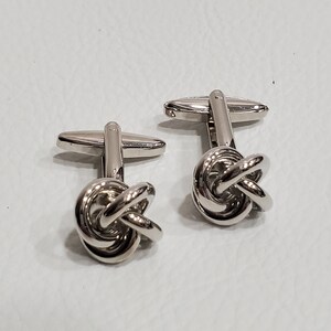 Könnte beinhalten: Ein Paar silberne Manschettenknöpfe mit einem Knotendesign. Die Manschettenknöpfe sind aus Metall und haben eine polierte Oberfläche.