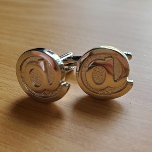 Puede incluir: Un par de gemelos de plata con un diseño de símbolo de arroba.