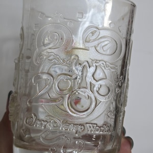 May include: Clear glass tumbler with embossed design. The design includes the number "2016" and the words "Disney World". The glass has a cylindrical shape and is likely used for beverages.