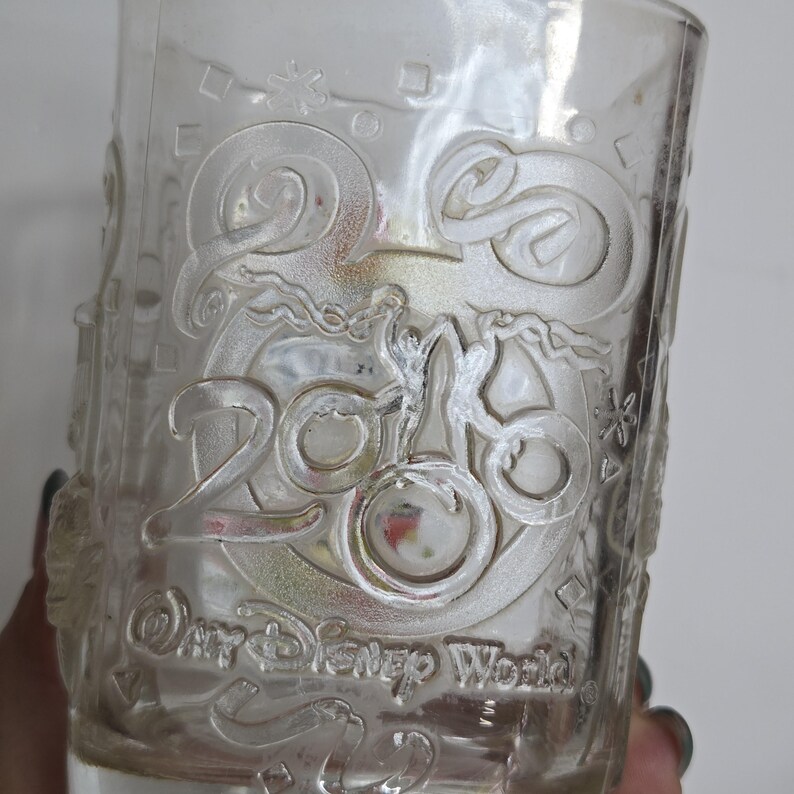 May include: Clear glass tumbler with embossed design. The design includes the number "2016" and the words "Disney World". The glass has a cylindrical shape and is likely used for beverages.