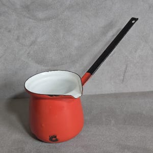 May include: A small, red enamelware pot with a long, black handle. The interior is white, and the rim is also white. The pot has a spout for easy pouring. A classic kitchenware item.