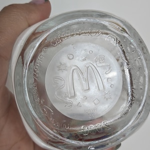 May include: Clear glass cup with the McDonald's logo on the bottom. The logo is a large, transparent "M" shape, surrounded by various shapes and the text "CELEBRATION". The cup is held by a hand.