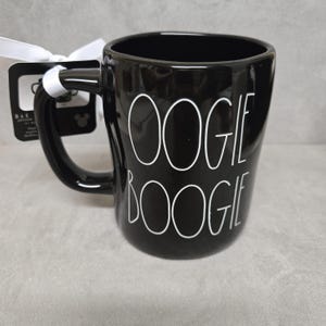 May include: A glossy black ceramic mug featuring the white text OOGIE BOOGIE printed in a tall, thin font. A white ribbon is tied to the handle, securing a black rectangular product tag with white text and a small Mickey Mouse icon.