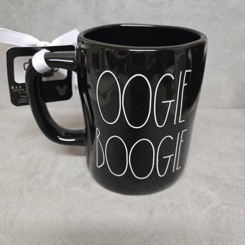 May include: A glossy black ceramic mug featuring the white text OOGIE BOOGIE printed in a tall, thin font. A white ribbon is tied to the handle, securing a black rectangular product tag with white text and a small Mickey Mouse icon.