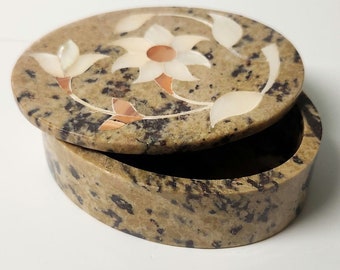 May include: A small oval-shaped trinket box made of brown stone with a floral design. The lid is open and features a white flower with a pink center and leaves made of mother of pearl.