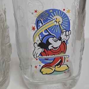 May include: Clear glass tumbler featuring a colorful graphic of Mickey Mouse in a sorcerer's outfit, with a blue and yellow design. The design includes stars and fireworks. The number "2000" is printed at the bottom of the glass.