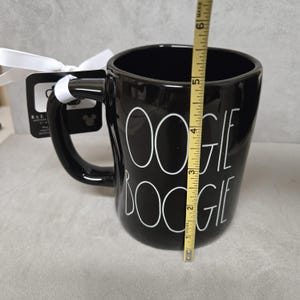 May include: A black ceramic mug with the white text OOGIE BOOGIE printed on the front. A yellow measuring tape is positioned vertically against the mug, showing a height of approximately 6 inches. A white ribbon and a black product tag are attached.