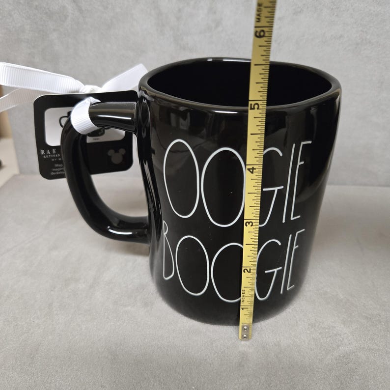 May include: A black ceramic mug with the white text OOGIE BOOGIE printed on the front. A yellow measuring tape is positioned vertically against the mug, showing a height of approximately 6 inches. A white ribbon and a black product tag are attached.