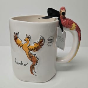 Puede incluir: Taza de cerámica blanca con una ilustración de fénix y la palabra "fawkes" impresa en la parte delantera. La taza tiene un asa de loro rojo y amarillo. La taza es de doble cara y tiene una etiqueta que dice "Rae Dunn"
