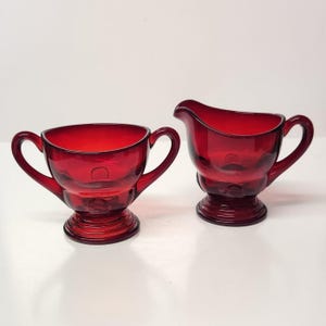 May include: A ruby red glass sugar bowl and creamer set. The sugar bowl has two handles and a pedestal base. The creamer has a spout and a matching pedestal base. Both pieces are made of transparent glass.
