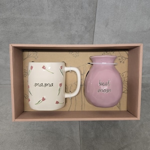 Puede incluir: Un juego de regalo con una taza de cerámica blanca con la palabra "mama" y detalles florales, y un jarrón rosa con la inscripción "best mom". Los artículos se presentan en una caja de regalo rosa, ideal para el Día de la Madre.