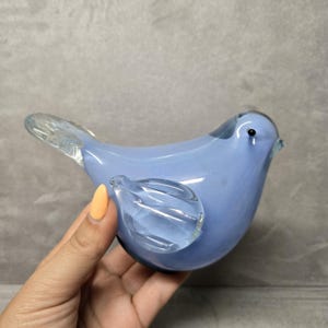 Puede incluir: Figurita de pájaro de cristal azul claro con alas y cola de cristal transparente. El pájaro tiene un pequeño ojo negro y un pico ligeramente curvado. La figura es lisa y brillante.