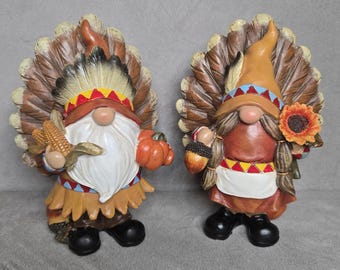 May include: Two gnome figurines wearing feathered headdresses. The left gnome has a white beard, holds a corn cob and an orange pumpkin. The right gnome has braided hair, holds an acorn and a sunflower. Both wear brown outfits with black boots.