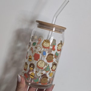 May include: A clear glass drinking cup with a bamboo lid and a clear glass straw. The cup is decorated with colorful cartoon character stickers. The stickers include images of Charlie Brown, Snoopy, and other Peanuts characters. The cup is designed for cold beverages.