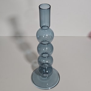 May include: A blue glass candle holder with a cylindrical top, three spherical sections, and a circular base. The candle holder is made of transparent glass and has a modern, minimalist design. The height is approximately 10 inches.