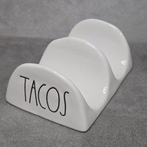 以下が含まれることがあります： 3つのスロットが付いた白いセラミックタコスホルダー。「TACOS」の文字が前面に黒の様式化された文字で書かれています。ホルダーはタコスを直立に保つように設計されており、キッチンやダイニングでの使用に適しています。