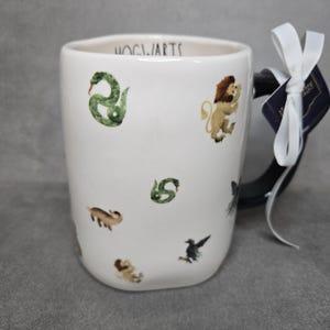 May include: White ceramic mug with black handle, featuring illustrations of a snake, lion, badger, raven, and text that reads "HOGWARTS". A white ribbon and tag are attached to the handle.