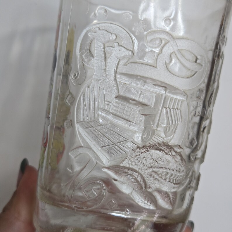 May include: Clear glass with an embossed design featuring a giraffe, a trolley, and other decorative elements. The glass has a slightly tapered shape and is partially filled, with the design visible on the exterior.