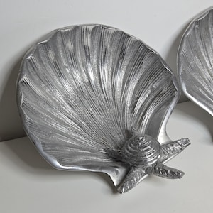 May include: Two silver seashell-shaped decorative trays. Each tray has a detailed shell design with a starfish and a small turtle accent. The trays are made of a metallic material, suitable for coastal or beach-themed decor.