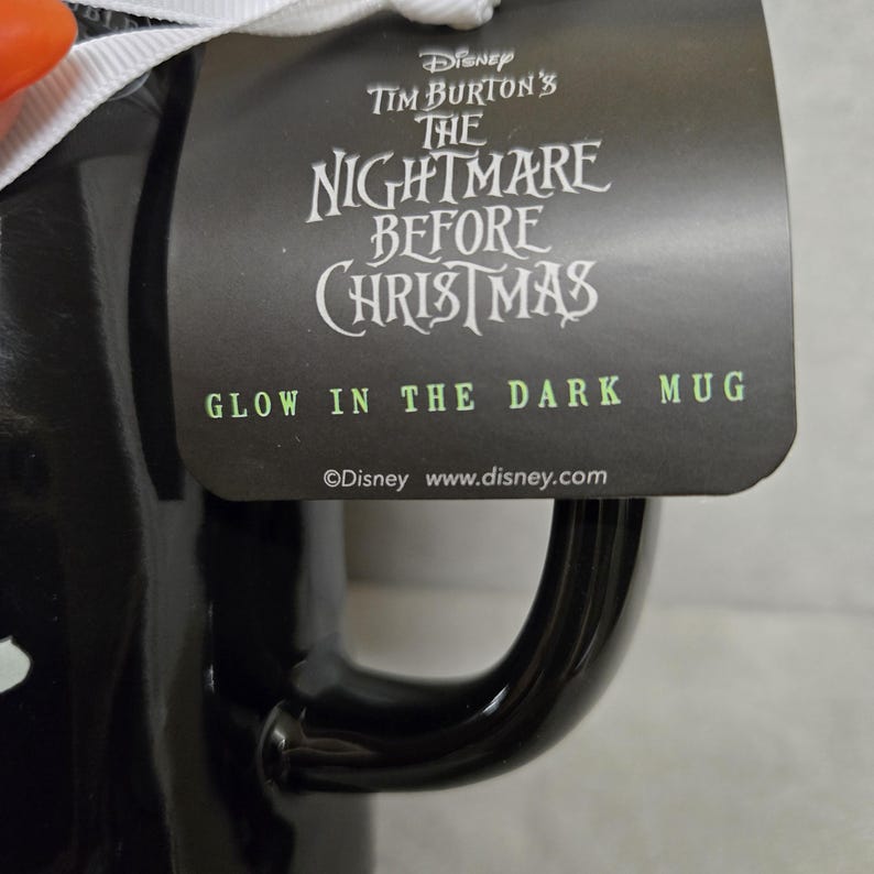 May include: A black ceramic mug featuring a tag that reads Disney Tim Burton's The Nightmare Before Christmas Glow In The Dark Mug. The tag is attached with a white ribbon and includes the Disney logo and website address at the bottom.