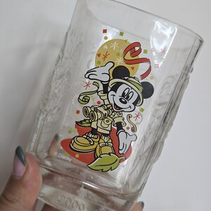 May include: Clear glass featuring a cartoon Mickey Mouse in safari attire. The design includes a yellow background, red ribbon, and green confetti. The glass has a textured pattern and the Disney logo.