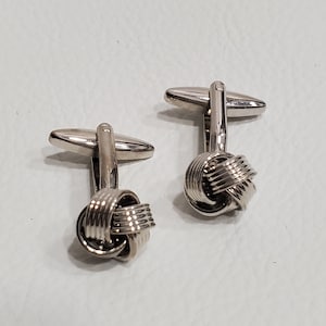 Puede incluir: Un par de gemelos de plata con un diseño de nudo. Los gemelos están hechos de metal y tienen un acabado pulido.