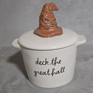 Puede incluir: Un recipiente de cerámica blanco con tapa, con un Sombrero Seleccionador marrón. El recipiente tiene dos asas pequeñas y las palabras "deck the great hall" en escritura negra. Un artículo decorativo.