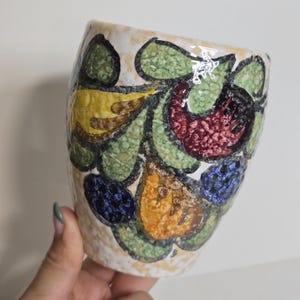 May include: A ceramic vase with a cream-colored speckled background. The vase is decorated with a colorful floral pattern featuring green, yellow, red, blue, and orange shapes. The vase has a slightly curved shape.