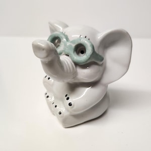 May include: White ceramic elephant figurine wearing green glasses. The elephant is sitting with its trunk raised and has black dots on its legs.