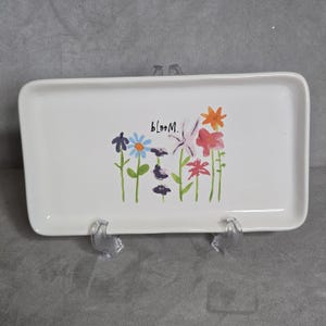Puede incluir: Una bandeja de cerámica rectangular blanca con esquinas redondeadas. La bandeja está decorada con un diseño floral colorido y la palabra "bloom". Las flores son en tonos de azul, naranja, rosa y morado. La bandeja se muestra sobre un soporte de plástico transparente.