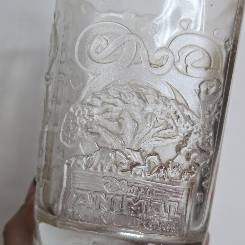 May include: Clear glass tumbler with embossed design featuring a tree, animals, and the text "Disney's Animal Kingdom". The glass has a slightly flared shape and is partially filled.