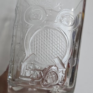 May include: Clear glass tumbler with embossed design. Features a tennis racket, fireworks, and the text "EPCOT". The design is surrounded by stars and other decorative elements. A collectible item.