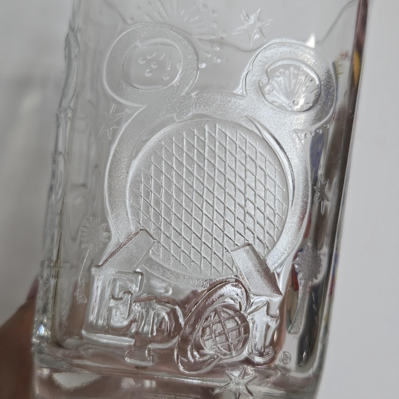 May include: Clear glass tumbler with embossed design. Features a tennis racket, fireworks, and the text "EPCOT". The design is surrounded by stars and other decorative elements. A collectible item.