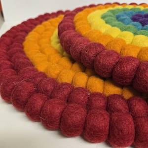 May include: A felted wool trivet in a rainbow design. The trivet features concentric circles of red, orange, yellow, green, blue, and purple. The trivet is made of small, round felt balls, creating a textured surface. The trivet is designed to protect surfaces from heat.