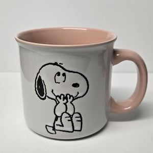 May include: A white ceramic mug with a pink rim and handle. The mug features a black and white illustration of Snoopy sitting with his paws in his lap.