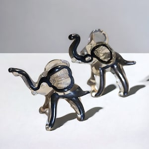 May include: Two clear glass elephant figurines with black painted details. The elephants are standing on a white surface.