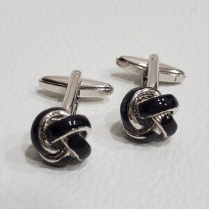 Könnte beinhalten: Ein Paar silberne Manschettenknöpfe mit schwarzen Knoten aus Emaille.