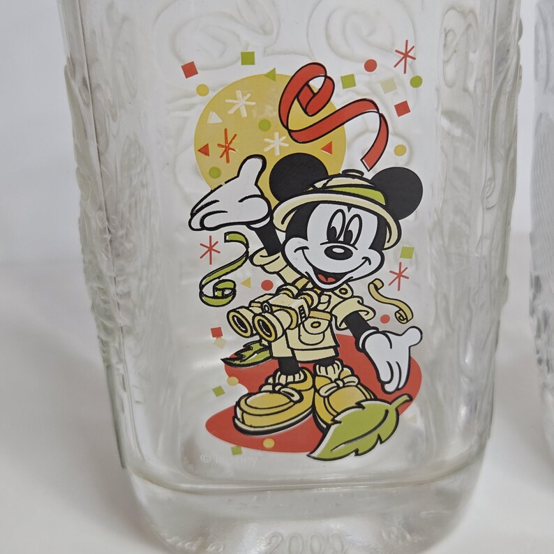 May include: Clear glass tumbler featuring a cartoon image of Mickey Mouse in safari attire. The design includes a yellow circle with red and green accents, and the year 2005 is printed at the bottom. The glass has a textured pattern.