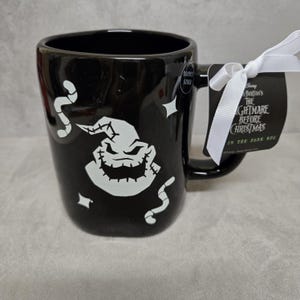 May include: A black ceramic mug featuring a white graphic of the character Oogie Boogie from The Nightmare Before Christmas, surrounded by small white worms and stars. A white ribbon attaches a tag with the movie title and Glow in the Dark text.