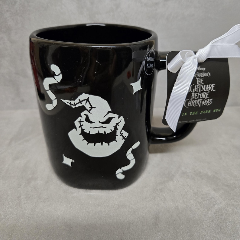 May include: A black ceramic mug featuring a white graphic of the character Oogie Boogie from The Nightmare Before Christmas, surrounded by small white worms and stars. A white ribbon attaches a tag with the movie title and Glow in the Dark text.