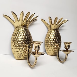 Pode incluir: Duas arandelas douradas em forma de abacaxi. As arandelas têm um padrão texturizado, semelhante a favos de mel, no corpo e um castiçal. Os topos de abacaxi têm um design espinhoso em forma de folha. Os castiçais são curvos.