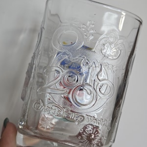 May include: Clear glass tumbler with embossed "2000" and "Walt Disney World" text. The glass features a decorative design with floral and Mickey Mouse head accents. The glass is partially filled with colorful items.