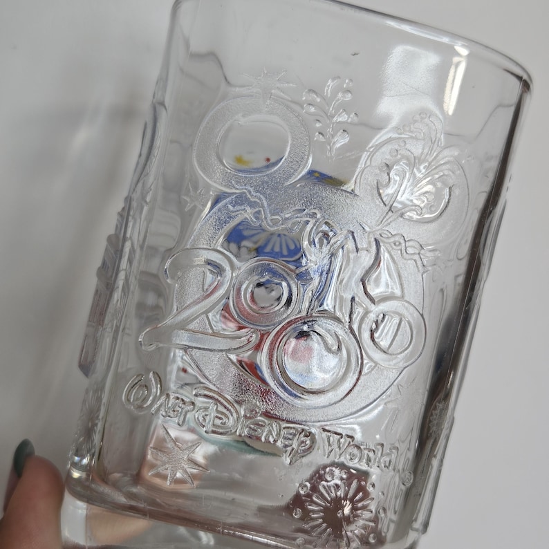 May include: Clear glass tumbler with embossed "2000" and "Walt Disney World" text. The glass features a decorative design with floral and Mickey Mouse head accents. The glass is partially filled with colorful items.